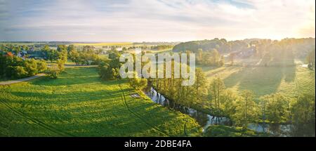 Frühling Morgen Luft ländlichen Panorama. Sonnenaufgang über grün und gelb blühenden Colza Feldern. Nebelwolken und kleiner Fluss mit Bäumen am Flussufer. Bela Stockfoto