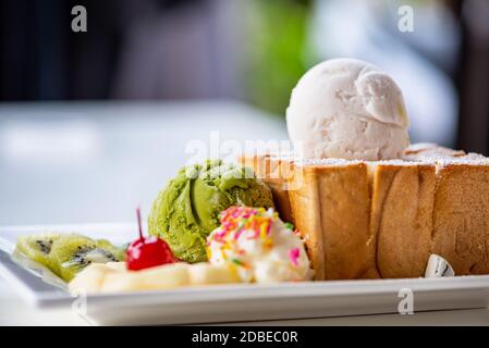 Closeup Honig Toast Brot serviert mit grünem Tee und Kokosmilch Eiskugel, dekorieren mit Kirsche, Apfel, Kiwi-Obst und Sahne Zucker Topping spr Stockfoto