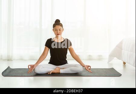 Frauen mittleren Alters üben Yoga in Easy Seat Pose oder Sukhasana mit Mudra. Meditation mit Yoga im weißen Schlafzimmer nach dem Aufwachen am Morgen. Stockfoto