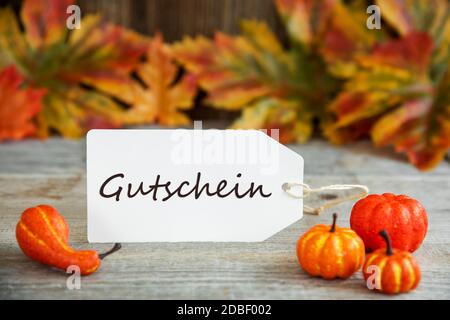 Weißes Etikett Mit Deutschem Text Gutschein Bedeutet Gutschein. Holzhintergrund Mit Herbstdeko Wie Kürbis Und Blätter Stockfoto