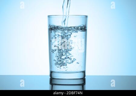 Wasserstrahl füllt ein Glas steht auf einem glänzenden Tisch mit Reflexion. Wasserspritzer mit Blasen. Kaltblaue getönte Vignette. Stockfoto