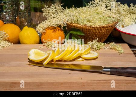 Auf einem Schneidebrett liegt ein Küchenmesser und eine in Scheiben geschnittene Bio-Zitrone, im Hintergrund sieht man Orangen, Zitronen und andere Zutaten Stockfoto