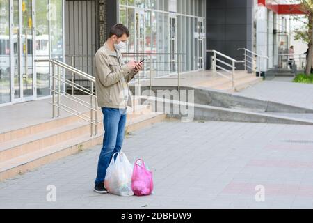 Ein Mann in einer medizinischen Maske und ein Einkauf hielten auf einer leeren Straße an, um einen Anruf anzunehmen Stockfoto