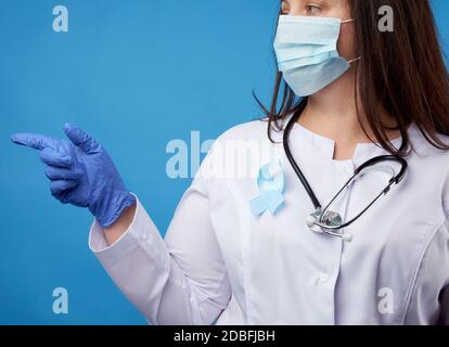 Ärztin in einer sterilen Einweg-Maske und einem weißen Mantel steht auf einem blauen Hintergrund und zeigt mit dem Zeigefinger zur Seite, ein blauer Seidenrippen Stockfoto