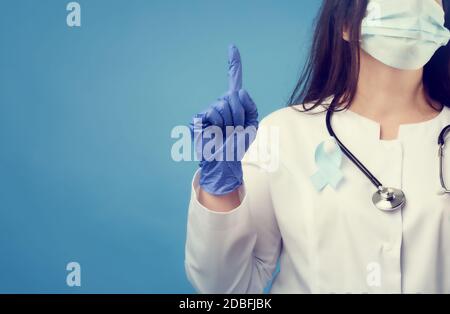 Ärztin in einer sterilen Einweg-Maske und einem weißen Mantel steht auf einem blauen Hintergrund und zeigt mit dem Zeigefinger nach oben, ein blaues Seidenband in die Stockfoto