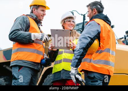 Drei Arbeiter in einem Steinbruch diskutieren vor schweren Maschinen, um Plan zu betrachten Stockfoto