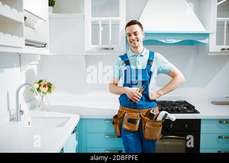 Fröhlicher männlicher Klempner in Uniform Posen in der Küche. Handwerker mit Werkzeugtasche Reparatur Waschbecken, Sanitär-Service zu Hause Stockfoto
