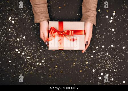Geschenkbox mit roter Schleife in den Händen der Frau. Dunkler Hintergrund mit Funkeln. Stockfoto