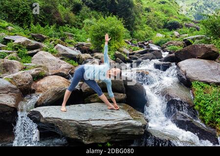 Frau, die Ashtanga Vinyasa Yoga asana Utthita trikonasana - erweitert Triangle Pose im Freien am Wasserfall im Himalaya Stockfoto