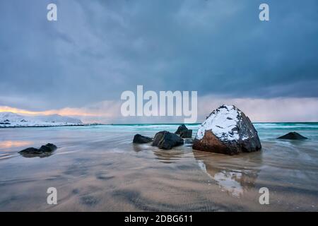 Felsen mit Schnee bedeckt am norwegischen Meer Strand in Fjord bei stürmischem Wetter mit Wolken. Skagsanden Strand, Flakstad, Lofoten Inseln, Norwegen. Lange Ausr Stockfoto