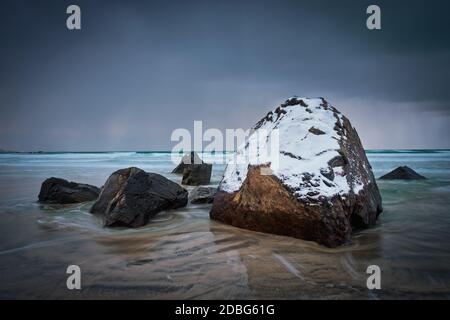 Felsen mit Schnee bedeckt am norwegischen Meer Strand in Fjord bei stürmischem Wetter mit Wolken. Skagsanden Strand, Flakstad, Lofoten Inseln, Norwegen. Lange Ausr Stockfoto