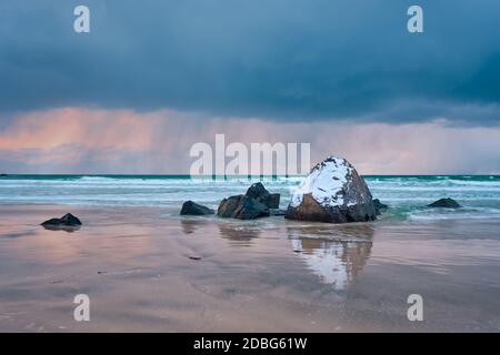 Felsen mit Schnee bedeckt am norwegischen Meer Strand in Fjord bei stürmischem Wetter mit Wolken. Skagsanden Strand, Flakstad, Lofoten Inseln, Norwegen. Lange Ausr Stockfoto