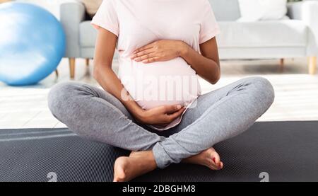 Unkenntlich schwarze schwangere Frau sitzt in Lotuspose auf Yoga-Matte und umarmt ihren großen Bauch zu Hause, Panorama Stockfoto