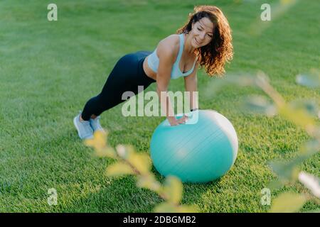 Zufrieden schlanke Frau lehnt an fit Ball, in aktiver Kleidung gekleidet, hat Gymnastik-Übungen auf grünem Rasen, genießt Sport und Lieblings-Aktivität, führt gesund Stockfoto