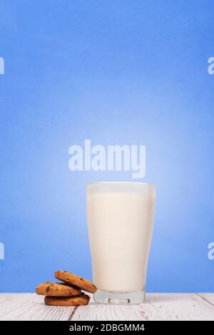 Chocolate Chip Cookies sitzen neben einem vollen Glas Milch auf einer Holzfläche gegen eine blaue Strukturwand Mit Platz für Text und Platz für Text Stockfoto