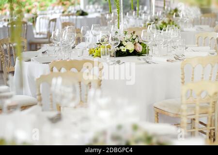 Formelle Tischaufstellung in einem Hochzeitslokal mit stilvollen weißen Stühlen, eleganten Gläsern und Silberbesteck um florale Mittelstücke mit selektivem Fokus Stockfoto