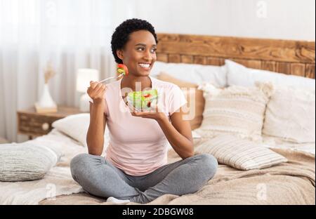 Gesunde Ernährung zur Gewichtsabnahme Konzept. Glückliche schwarze Frau essen leckeren Gemüsesalat auf dem Bett zu Hause, Platz leer Stockfoto