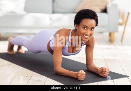 In voller Länge Porträt von lächelnden schwarzen Frau in Sportswear tun Ellenbogen Plank auf Sportmatte zu Hause Stockfoto