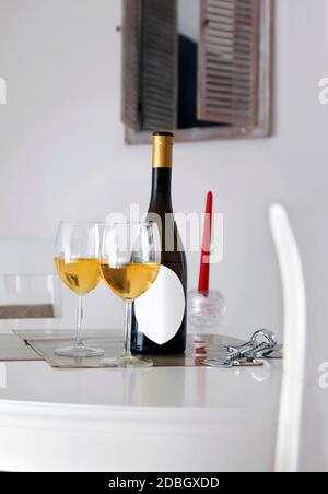 Flasche Wein mit weißem Etikett für Beschriftung, Öffner, zwei Gläser Weißwein auf dem Tisch und Fenster im Hintergrund Stockfoto