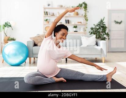 Gesundes, gesundes Schwangerschaftskonzept. Glückliche schwarze werdende Frau streckt ihr Bein, Yoga auf Matte zu Hause üben Stockfoto