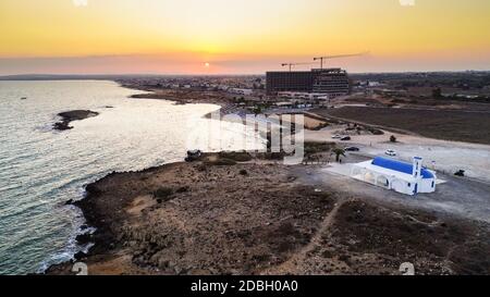 Blick auf den Sonnenuntergang an der Küste und die traditionelle weiße, gewaschene Kapelle mit blauen Türen am Strand von Agia Thekla, Ayia Napa, Famagusta, Zypern von A Stockfoto