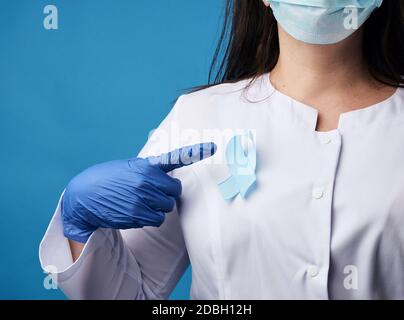 Ärztin in einer sterilen Einweg-Maske und einem weißen Mantel steht auf blauem Hintergrund und zeigt mit seinem Zeigefinger, blaues Seidenband in den Schap Stockfoto