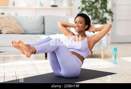 Sport in Quarantäne. Attraktive schwarze Frau macht Fitness auf Sportmatte zu Hause, die Ausübung ihrer bauchmuskeln Stockfoto