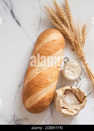 British White Bloomer oder europäischen Sauerteig Baton Laib Brot auf weißem Marmor Hintergrund. Frisches Brot und Glas mit Sauerteig Starter, Mehl Stockfoto