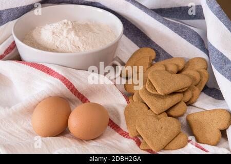 Gourmet-Nahaufnahme mit einer Schüssel voller Mehl, herzförmigen Keksen und zwei Eiern in weißen Küchentüchern mit roten und blauen Rand, auf einem Holzbrett Stockfoto