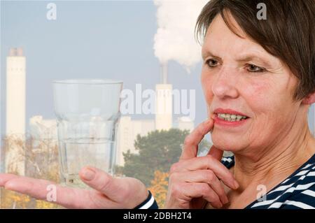Seitliche Kopf-und-Schultern-Ansicht einer älteren Frau mit einer skeptischen Ansicht eines halb gefüllten Glases mit Wasser auf der rechten Seite Hand, die gegen eine Industria steht Stockfoto
