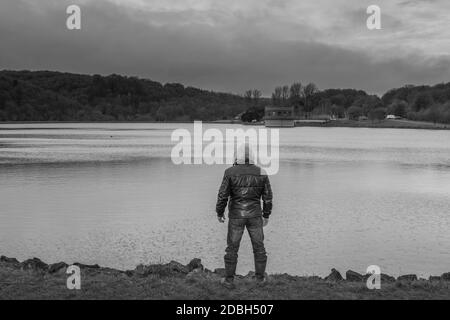 Schwarz-weiße Rückansicht eines bedrohlichen Mannes mit einer Kapuze über dem Wasser mit launischen Wolken darüber. Stockfoto