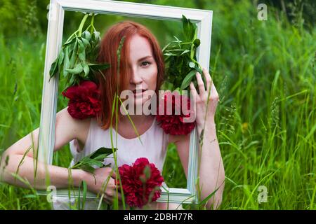 Rotschopf kaukasische Frau hält einen Bilderrahmen mit roten Pfingstrosen Blumen geschmückt. Hochzeitsfotozone Konzept. Stockfoto