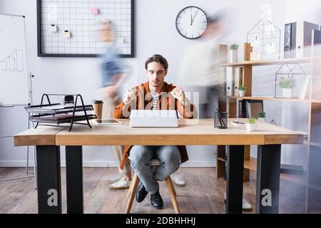 Der junge Manager sitzt mit geschlossenen Augen und geballten Fäusten am Schreibtisch im offenen Büro, Bewegungsunschärfen Stockfoto