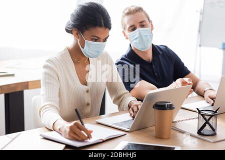 afroamerikanische Geschäftsfrau tippt auf Laptop ein Schreiben in Notebook In der Nähe Kollege in medizinische Maske auf verschwommenem Hintergrund Stockfoto