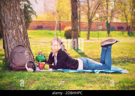 Schönes lächelndes junges Mädchen, das auf dem grünen Gras im Park liegt und eine Münze in ein grünes Sparschwein steckt. Sparkonzept und Geldkonzept. Schüler speichern c Stockfoto