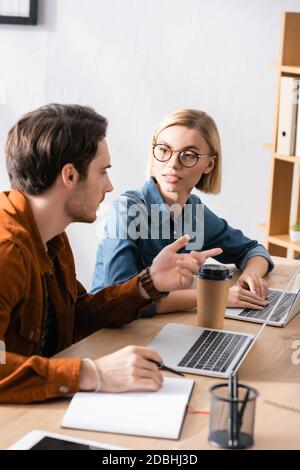 Blonde Frau in einer Brille, die die gestikulierenden Kollegen ansieht, während sie auf unscharfem Hintergrund am Schreibtisch mit Laptops im Büro sitzt Stockfoto