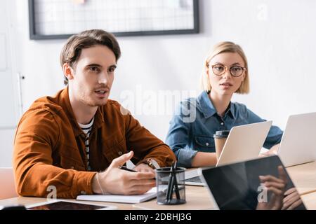 Blonde Frau sitzt am Arbeitsplatz in der Nähe Mann reden und gestikulieren Im Büro auf unscharfem Hintergrund Stockfoto