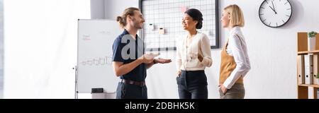 Lächelnde, multikulturelle Büromitarbeiter sprechen, während sie neben dem Maschenorganisator mit leeren Aufklebern und Flipchart, Banner stehen Stockfoto