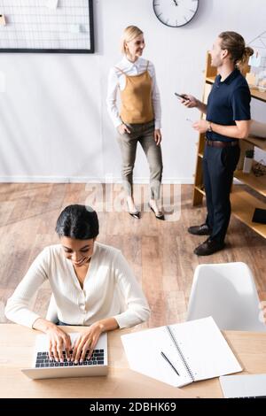 Overhead-Ansicht der glücklichen afroamerikanischen Frau tippen auf Laptop Während sie am Schreibtisch sitzen und Büroangestellte im Hintergrund sprechen Stockfoto