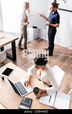 Ansicht der afroamerikanischen Frau mit Laptop während des Sitzens Am Tisch mit Büroangestellten, die auf dem Hintergrund sprechen Stockfoto