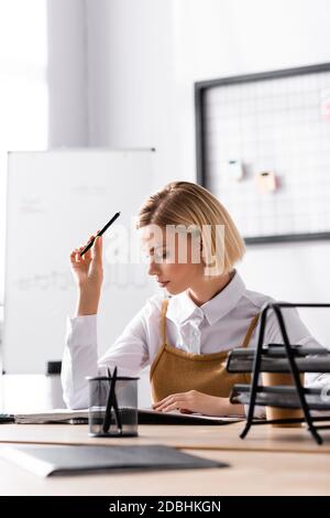 Fokussierte blonde Frau mit Stift Blick auf Notizblock während des Sitzens Am Schreibtisch im Büro auf unscharfem Hintergrund Stockfoto