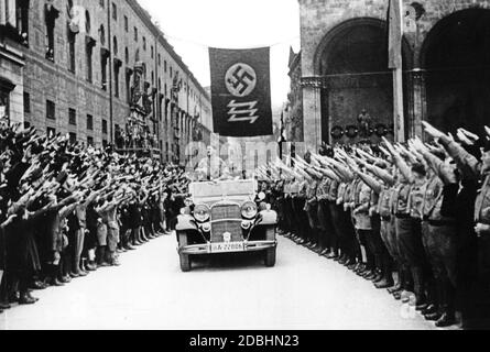 "Adolf Hitler und die NSDAP feiern die Erreichung ihrer Ziele am 9. November 1933, 10 Jahre nach dem gescheiterten Putsch. Hitler war in diesem Jahr Reichskanzler geworden. An der Stelle unter der Flagge mit Hakenkreuz und Wolfsangel, die im Hintergrund zu sehen ist, wurde eine Gedenktafel zu Ehren der Märtyrer vom 9. November 1923 angebracht, die nur durch das Angrüßen mit dem Nazi-Gruß übergeben werden konnte. Ab diesem Zeitpunkt wurde der 9. November zum nationalsozialistischen Feiertag, in dessen Rahmen der traditionelle marsch vom Bürgerbraeukeller am Wiener Platz über die Ludwigsbrücke stattfand Stockfoto