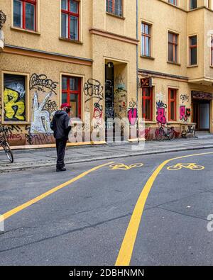 Ein älterer Mann mit Gesichtsmaske geht entlang der Straße neben der Fahrradstraße außerhalb der Graffiti-bedeckten Außenfassade der Schmittz Bar, Mitte, Berlin Stockfoto