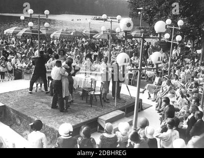 "An einem schönen Sommertag 1931 fand am Krumme Lanke See in Berlin-Zehlendorf ein Filmfestival statt. Das Publikum spielt und ihre Emotionen und Reaktionen werden von einer Tonfilmkamera aufgenommen, die auf der Bühne steht. Auf zwei Sonnenschirmen wird Milch mit den Slogans 'Milch macht stark' und 'frisch durch frische Milch' beworben. Auf der Bühne steht eine Kamera, die auf die Kamera des Fotografen zeigt." Stockfoto