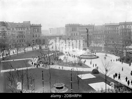 Das Foto zeigt den Belle Allliance Platz in Berlin um 1910 mit der Friedenssäule in der Mitte und einem Blick in Richtung Hallesches Tor mit der Hochbahn (im Hintergrund). Es gibt viele Wanderer im Kreisverkehr, und am Rand gibt es zwei Straßenbahnen. Die Darmstaedter Nationalbank befindet sich im Gebäude ganz links. Stockfoto