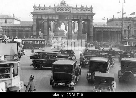 Das Bild zeigt das Brandenburger Tor in Berlin, das anlässlich der Hindenburg-Feier 1928 festlich geschmückt und mit Fahnen aufgehängt wurde. Davor fahren Autos, Busse und Straßenbahnen, während Polizisten versuchen, den Verkehr zu regulieren. Stockfoto