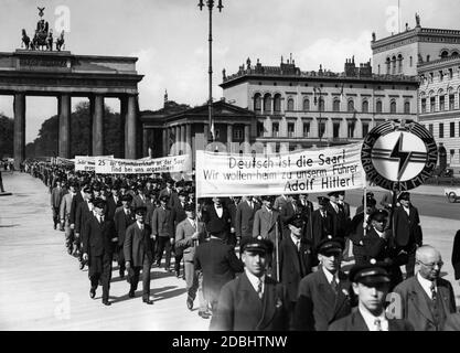 "Etwa zweitausend Saardeutsche, die anlässlich der Funkausstellung nach Berlin kamen, marschieren auf dem Weg zur Reichskanzlei durch das Brandenburger Tor. Vorne steht der Standard einer Delegation des VDA aus dem Landkreis Saarbrücken in Malstatt. Dahinter stehen mehrere Banner mit der Aufschrift: "Die Saar ist deutsch! Wir wollen nach Hause zu unserem Führer Adolf Hitler'' und ''25% des gesamten Publikums in der Saar sind um uns herum organisiert'''. Stockfoto