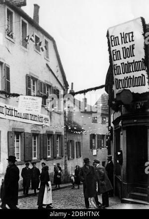 "Die Altneugasse von Saarbrücken ist feierlich mit Hakenkreuzfahnen, kaiserlichen Fahnen und Bannern geschmückt, um den ersten Jahrestag der "Machtausweitung" zu feiern. Links: 'Saar strebt treu nach dem neuen Reich 1934'. In der Mitte ein Text '' an den Führer!''. Rechts der Slogan: ''One Heart. Eine Nation. Ein Vaterland. Deutschland''.' Stockfoto