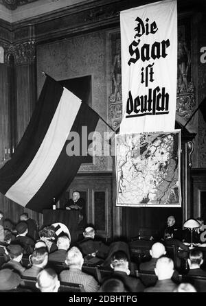 "Kommerzienrat Hermann Roechling spricht auf dem Rednerpult bei einer Saar-Kundgebung der Deutschen Weltwirtschaftsgesellschaft am Oberverwaltungsgericht in der Hardenbergstraße in Saarbrücken. An der Wand, neben der Reichsflagge, befindet sich eine Karte von 'Saar- und Mosellandschaft, Pfalz und Rheinhessen' und ein Banner mit der Aufschrift ''die Saar ist deutsch'''. Stockfoto