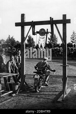 Ein Mitglied der Motor HJ bei einem Fahrtest während des Sportfestivals der Hitlerjugend auf dem Westend Sportplatz in Berlin. Er fährt unter einem Wassereimer. Das Publikum ist im Hintergrund. Auf der linken Seite des Bildes sind andere Mitglieder des HJ lachend. Er fährt ein Motorrad der Marke DKW. Stockfoto